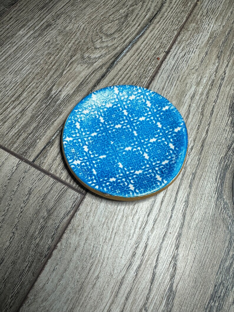 May include: A round coaster with a blue and white patterned design. The coaster has a gold-colored edge. The pattern is a repeating geometric design. The coaster is sitting on a wooden surface.