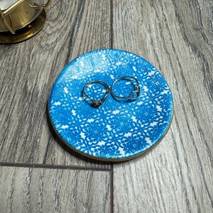 May include: A small, round, blue and white patterned ceramic ring dish with a gold rim. Two silver rings are on the dish. The dish is on a wooden surface. A gold candle holder is in the background.