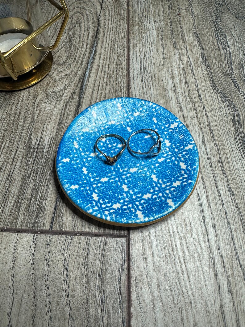 May include: A small, round, blue and white patterned ceramic ring dish with a gold rim. Two silver rings are on the dish. The dish is on a wooden surface. A gold candle holder is in the background.