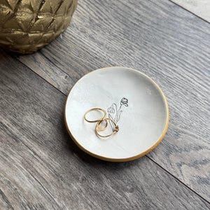 May include: A small, round, white ceramic ring dish with a gold rim. The dish features a delicate black floral design and holds two gold rings. The background is a wooden surface, and a gold decorative object is partially visible.
