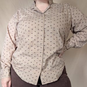 May include: Tan button-down shirt with a small, dark floral pattern. The long-sleeved shirt has a single chest pocket and a classic collar. The shirt is paired with brown pants.