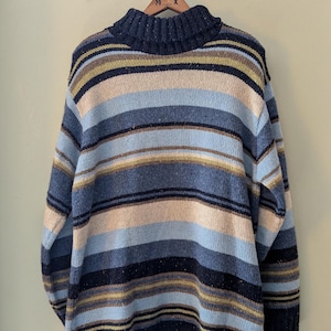 May include: A blue, beige, and brown striped sweater with a turtleneck. The sweater has horizontal stripes in various shades of blue, beige, and brown. The sweater is hanging on a wooden hanger.