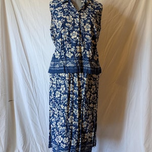 May include: A two-piece outfit featuring a sleeveless button-down top and a matching skirt. The ensemble is navy blue with a white floral pattern. The skirt is a midi length.