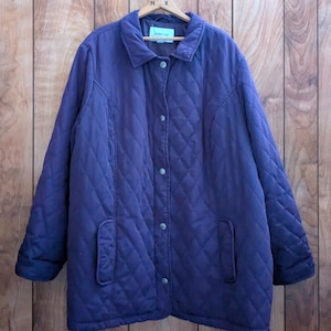 May include: A quilted, dark purple jacket with a button-up front and two front pockets. The jacket is hanging on a wooden hanger against a wooden background. The label on the jacket reads "avenue".