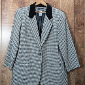 May include: A houndstooth blazer with a black velvet collar and a single-button closure. The jacket has two front pockets and is hanging on a wooden hanger. The blazer is black and white.