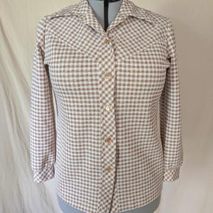 May include: A long-sleeve button-down shirt featuring a tan and white houndstooth pattern. The shirt has a pointed collar, a front button placket with five visible buttons, and cuffs at the end of each sleeve. The garment is displayed on a mannequin.