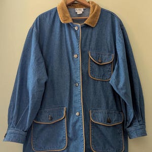 May include: A blue denim jacket featuring a tan corduroy collar, long sleeves with buttoned cuffs, and three front pockets with flaps. The jacket has a front button closure and tan piping trim along the edges and pocket flaps.