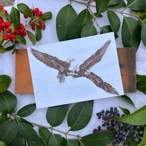 May include: A print featuring two eagles in flight, with outstretched wings and detailed feathering. The artwork is set against a light blue background, framed by green leaves, red berries, and wooden planks.