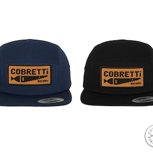 Può includere: Due cappellini da baseball a 5 pannelli con una toppa marrone che presenta il testo "COBRETTI WOOD WORKS" e una silhouette nera di un pesce con una lama da sega. Un cappello è blu navy e l'altro è nero.