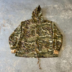 Puede incluir: Chaqueta de camuflaje con capucha, con un patrón de formas verdes, marrones y negras. La chaqueta tiene un cordón, múltiples bolsillos y puños color beige. La prenda está tendida sobre una superficie de hormigón.