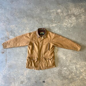 May include: A tan-coloured, vintage jacket with a brown leather collar and four front pockets. The jacket has a button-up closure and a small emblem on the collar. The jacket is laid flat on a concrete surface.