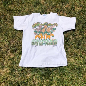 Puede incluir: Camiseta blanca con una ilustración de dibujos animados de jugadores de los Green Bay Packers en verde y amarillo. La camiseta tiene el texto "Magic and the Miracle" y "Green Bay Packers".