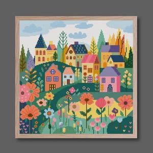 Könnte beinhalten: Ein gerahmtes Kreuzstich-Landschaftsbild zeigt ein farbenfrohes Dorf inmitten sanfter Hügel und lebendiger Blumen. Die Häuser haben Dächer in Blau, Rosa und Gelb. Das Kunstwerk ist vor einem hellblauen Himmel mit weißen Wolken angeordnet.