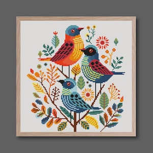 May include: A framed cross-stitch depicting three vibrant birds amidst foliage and flowers. The birds display blue, orange, and yellow hues with red and black details. The artwork is set against a white backdrop, framed in light wood.