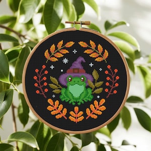 May include: Cross-stitch embroidery in a wooden hoop. The design features a green frog wearing a purple witch hat, surrounded by autumn foliage in orange, yellow, and green. The background is black.