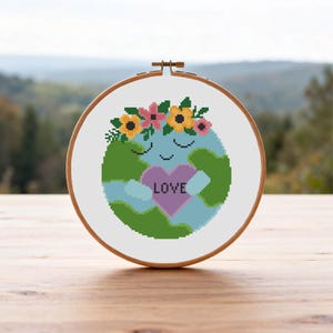 May include: A cross-stitch embroidery hoop featuring a smiling Earth with arms embracing a heart. The heart has the word "LOVE" on it. The Earth is wearing a floral crown. The background is a blurred view of trees.