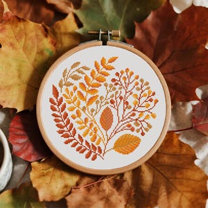 May include: A cross-stitch embroidery hoop with an autumnal design. The design features leaves and berries in orange, yellow, and brown hues. The embroidery is set on a white background and framed by a wooden hoop.