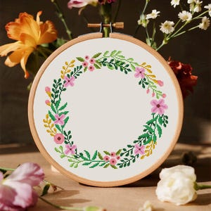 Könnte beinhalten: Ein runder Stickrahmen aus Holz mit einem weißen Stoffeinsatz, der ein Kreuzstichmuster eines Blumenkranzes zeigt. Der Kranz besteht aus rosa Blüten, grünen Blättern und gelben Zweigen, die kreisförmig auf dem Stoff angeordnet sind.
