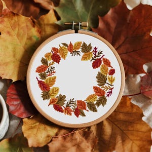 May include: A cross-stitch embroidery hoop with a wreath of autumn leaves in red, orange, yellow, and brown. The leaves are arranged in a circular pattern against a white background. The wooden hoop is set against a backdrop of fall foliage.