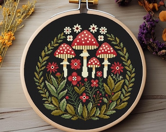 Op de afbeelding: Kruissteekborduurwerk in een houten hoepel met een paddestoelontwerp. De paddenstoelen hebben rode doppen met witte stippen, omringd door groene bladeren, rode bloemen en witte stervormige accenten. De achtergrond is zwart.