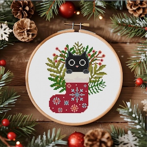 Peut inclure: Un projet de broderie au point de croix dans un tambour en bois rond représentant un chat noir sortant d'une chaussette de Noël rouge. La chaussette est ornée de flocons de neige bleu clair et blancs, entourée de branches de pin et de baies rouges.