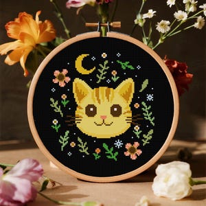May include: A cross-stitch embroidery hoop with a yellow tabby cat design on a black background. The cat has brown eyes and a smiling mouth. The design includes a crescent moon, flowers, and greenery. The hoop is made of wood and is surrounded by flowers.