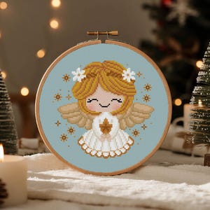 May include: A cross-stitch embroidery hoop featuring a smiling angel with golden hair, wings, and a star. The angel is dressed in white and beige, set against a light blue background. The wooden hoop is displayed on a white surface.
