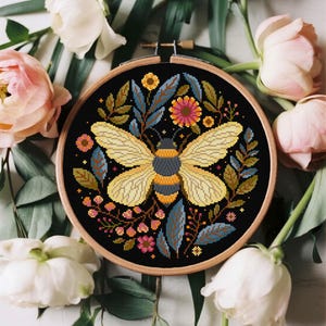 May include: A circular wooden embroidery hoop containing a cross-stitch pattern on black fabric. The design features a central yellow and grey bee surrounded by a symmetrical arrangement of colorful flowers, leaves, and small botanical elements.