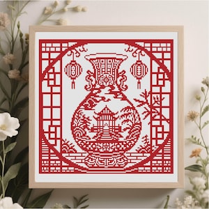 Pode incluir: Uma cruz bordada emoldurada com um design de vaso vermelho com uma pagoda, lanternas e padrões intrincados. A obra de arte é definida contra um fundo branco e emoldurada em madeira clara.