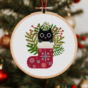 May include: A circular wooden embroidery hoop containing a cross-stitch pattern of a black cat peeking out of a red Christmas stocking. The stocking is decorated with light blue snowflakes and sits against a background of green pine branches and red berries.