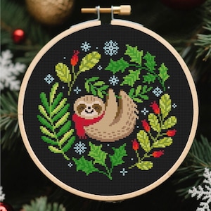 以下が含まれることがあります： 黒い布にクロスステッチの刺繍が施された円形の刺繍枠。赤いスカーフを巻いた薄茶色のナマケモノが中央に配置され、その周りを緑のヒイラギの葉、赤い実、水色の雪の結晶のアイコンがリースのように囲んでいるデザイン。