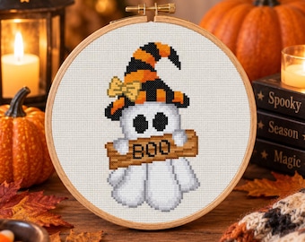 May include: A cross-stitch embroidery in a round wooden hoop featuring a white ghost wearing a black and orange striped witch hat with a small yellow bow. The ghost holds a brown wooden sign with the word BOO printed in black letters.
