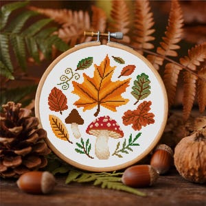 May include: A cross-stitch embroidery hoop featuring autumn leaves and a mushroom design. The leaves are in shades of orange, brown, and green, with a red-capped mushroom. The embroidery is set in a wooden hoop, with acorns and pinecones in the background.