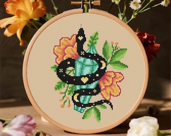 May include: A circular wooden embroidery hoop containing a cross-stitch pattern of a black snake coiled around a light blue crystal. The design features yellow heart-shaped markings on the snake, surrounded by yellow flowers and green leaves.