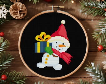 May include: A cross-stitch embroidery hoop featuring a snowman wearing a red hat and scarf, holding a blue gift with a yellow bow. The snowman is set against a black background, surrounded by Christmas decorations.