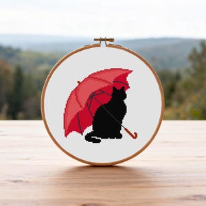 May include: A cross-stitch embroidery piece displayed in a round wooden hoop. The design features a solid black cat silhouette sitting under a large, vibrant red umbrella. The pattern is rendered in a pixelated style on a white fabric background.