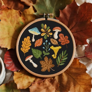 May include: A cross-stitch embroidery hoop featuring autumn leaves, mushrooms, and other fall foliage on a black background. The embroidery is set in a wooden hoop and surrounded by colourful autumn leaves.