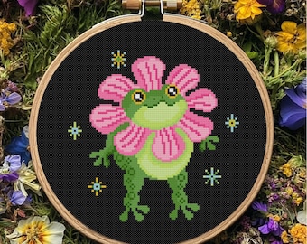May include: A circular wooden embroidery hoop containing black fabric with a cross-stitch design of a green frog wearing a large pink flower as a hat. The frog has a light green belly and four small, light blue star-shaped accents around it.