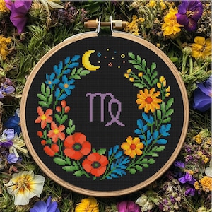 May include: A cross-stitch embroidery hoop featuring a black background with a floral wreath design. The wreath includes red poppies, blue and yellow flowers, and green leaves. The Virgo zodiac symbol is in the center, with a yellow crescent moon and stars above.