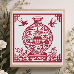 May include: A framed cross-stitch featuring a red and white design of a vase with a pagoda and birds. The artwork is in a wooden frame and surrounded by greenery.