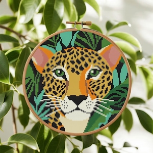 May include: A cross-stitch embroidery hoop featuring a leopard's face. The design includes a tan leopard with black spots, green eyes, and an orange nose. The background has green leaves and a teal border. The hoop is made of wood.