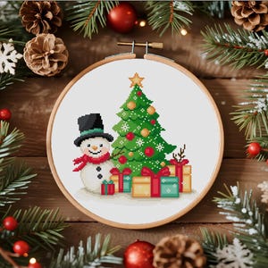 May include: A circular wooden embroidery hoop containing a cross-stitch pattern of a white snowman wearing a black top hat and a red scarf, standing next to a green Christmas tree decorated with gold ornaments and a yellow star, with gift boxes.