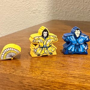 Puede incluir: Piezas de juego de madera que representan una figura amarilla con kimono, una figura azul y dos abanicos. Las figuras tienen rasgos faciales detallados y diseños de kimono. Los abanicos son amarillos y azules con detalles blancos.