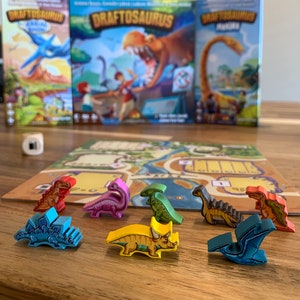 Puede incluir: Un juego de mesa llamado "Draftsaurus" con piezas de juego de dinosaurios de colores sobre una mesa de madera. Las piezas de juego son azules, verdes, amarillas, naranjas, rosas y rojas.