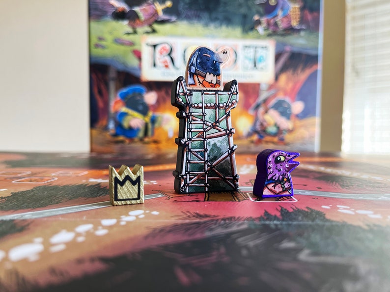 May include: A close-up of three game pieces from the board game "Root". The pieces are a wooden crown, a purple bird, and a tower with a green and brown design. The tower has a small, black figure on top.