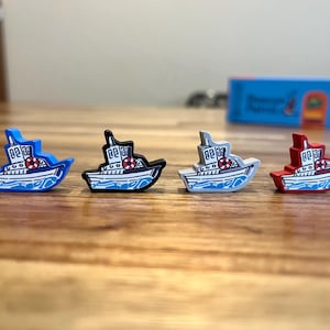 May include: Four wooden toy boats in different colors: blue, black, white, and red. Each boat has a white cabin with two windows and a red life preserver.