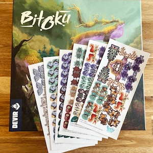 May include: A set of six sticker sheets for the board game Bitoku. The sheets feature colourful illustrations of various creatures, buildings, and other game elements. The stickers are printed on white paper and have a glossy finish.