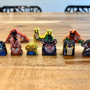 May include: A collection of nine small, flat, colorful wooden game tokens featuring illustrated animal characters and structures. The figures include a skunk, bat, frog, boar, cat, and bird, alongside three distinct building shapes in red, yellow, and orange.