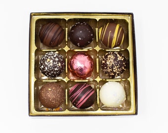 May include: A box of nine assorted chocolate truffles. The truffles are various colors and textures, including dark chocolate, white chocolate, and a pink foil-wrapped truffle. The box is gold-colored and sits on a white background.