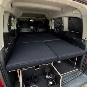 Puede incluir: Interior de furgoneta camperizada a medida con cama plegable y compartimentos de almacenamiento. La cama consta de dos cojines azul oscuro. El almacenamiento incluye tazas negras y objetos metálicos. El interior de la furgoneta es rojo.